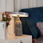 Grey marble Clérachic table lamp lit warm on a white dresser beside pink roses, in a bedroom with a dark blue bed.