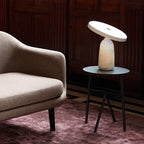 White marble Clérachic table lamp on a round metal side table, in a dark wood-panelled room with a beige armchair.