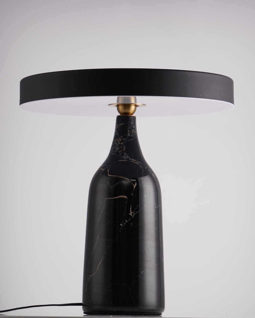 Black marble Clérachic table lamp on a dark table against a plain lavender-grey studio backdrop, shade held level.