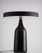 Black marble Clérachic table lamp on a dark table against a plain lavender-grey studio backdrop, shade held level.