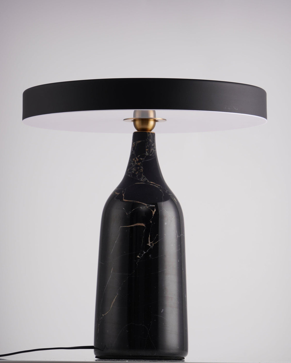 Black marble Clérachic table lamp on a dark table against a plain lavender-grey studio backdrop, shade held level.