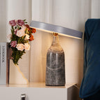 On a bedside table styled with pink roses and stacked books, this Clérachic lamp's charcoal marble base carries a tilted, glowing disc.