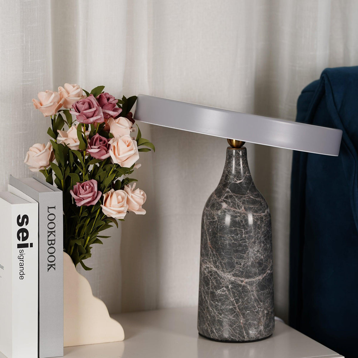 This Clérachic lamp sits unlit beside pink roses and books on a bedside table, its charcoal marble base and disc shade closed.