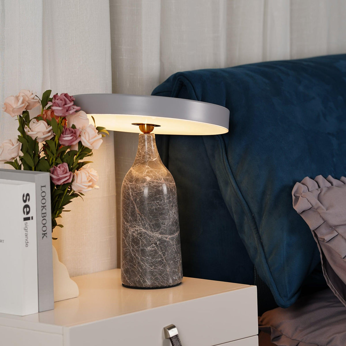Angled lower against the roses and books, this Clérachic lamp's charcoal marble base and tilted disc shade glow warm on the table.