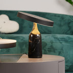 On a round side table against a deep teal sofa, this Clérachic lamp's black marble base carries a tilted disc shade glowing warm.