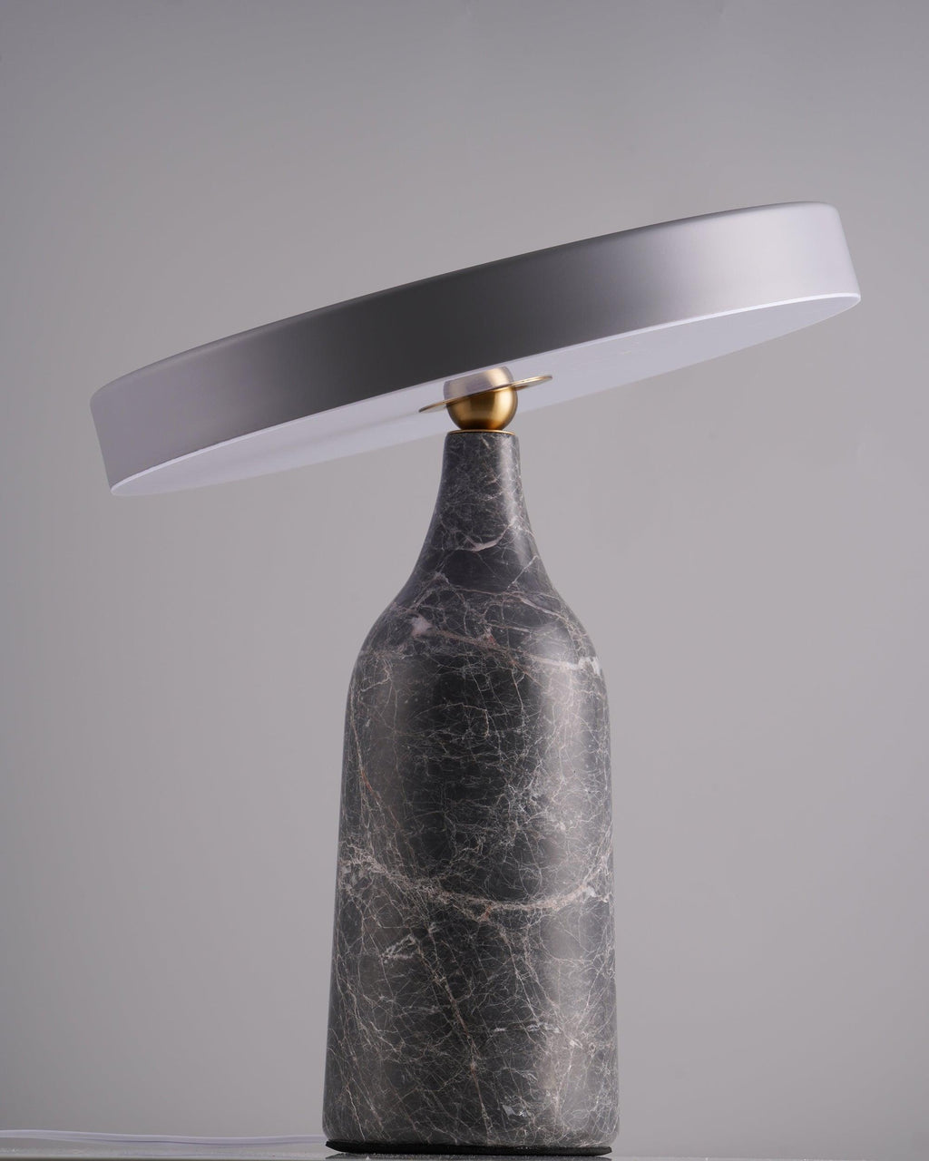 Against a plain grey studio backdrop, this Clérachic lamp's grey marble base carries its disc shade tilted at a shallow angle, switched off.