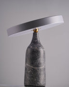 Against a plain grey studio backdrop, this Clérachic lamp's grey marble base carries its disc shade tilted at a shallow angle, switched off.