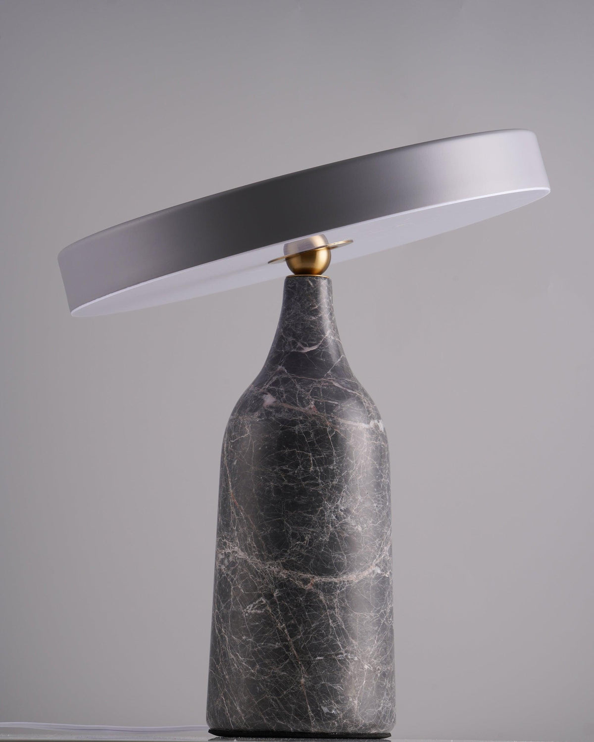 Against a plain grey studio backdrop, this Clérachic lamp's grey marble base carries its disc shade tilted at a shallow angle, switched off.