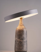 Lit against a soft grey gradient backdrop, this Clérachic lamp's grey marble base and tilted disc shade glow warm beneath the rim.