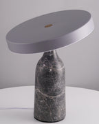 Photographed unlit against a plain grey backdrop, this Clérachic lamp's grey marble base supports its tilted disc, cord trailing to one side.