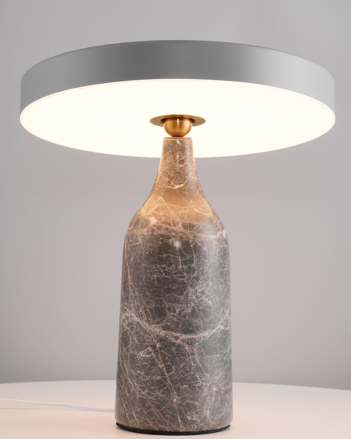 Against a soft grey studio backdrop, this Clérachic lamp's grey marble base carries its tilted disc shade lit with a pale glow.