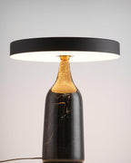 The black marble base of this Clérachic lamp carries a level disc shade glowing warm in a ring against a soft grey backdrop.