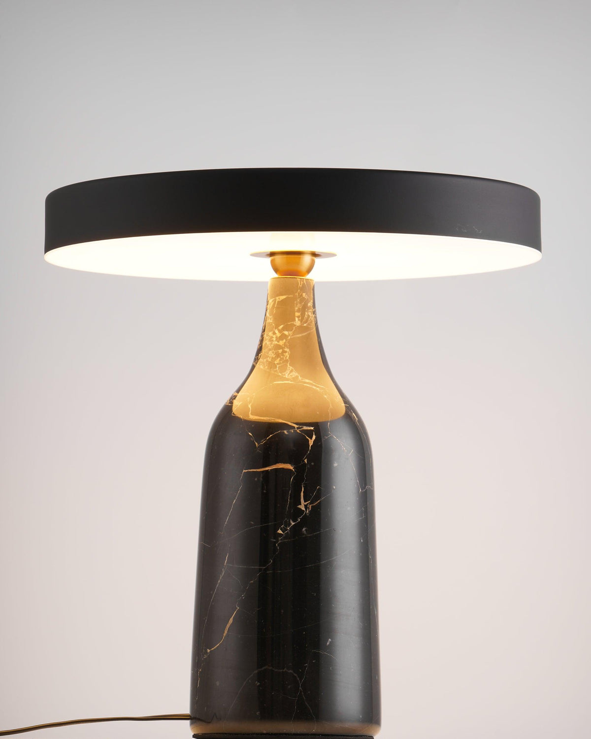 The black marble base of this Clérachic lamp carries a level disc shade glowing warm in a ring against a soft grey backdrop.