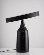 Tilted steeply on its black marble base, this Clérachic lamp's disc shade sits dark and unlit against a plain grey backdrop.