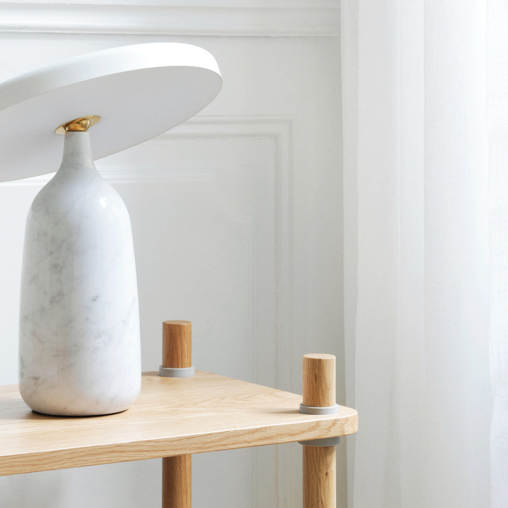 White marble Clérachic table lamp with its disc shade tilted steeply, close crop on the wooden two-tier shelf.