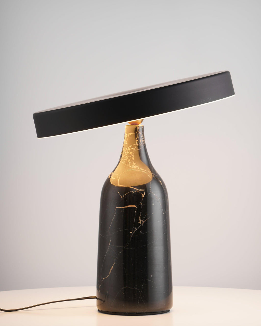 A black marble base veined with gold supports the steeply tilted, unlit disc shade of this Clérachic lamp on a white backdrop.