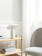 White marble Clérachic table lamp on the wooden shelf, photographed from a slightly wider angle near the window.