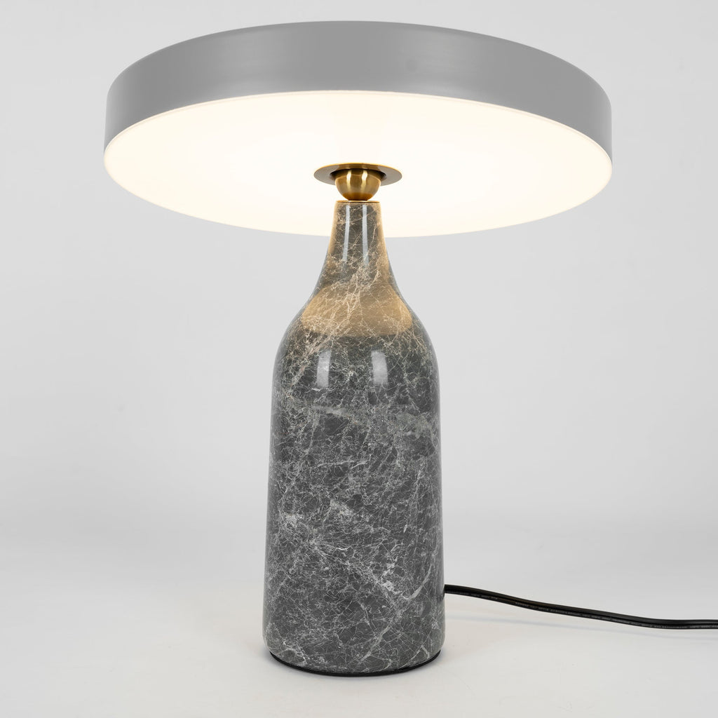 Grey marble Clérachic table lamp lit warm, its disc shade held level above the glowing base, tall studio crop.