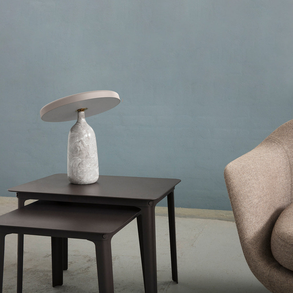 White marble Clérachic table lamp on dark nesting side tables beside a beige armchair, blue-grey wall behind.