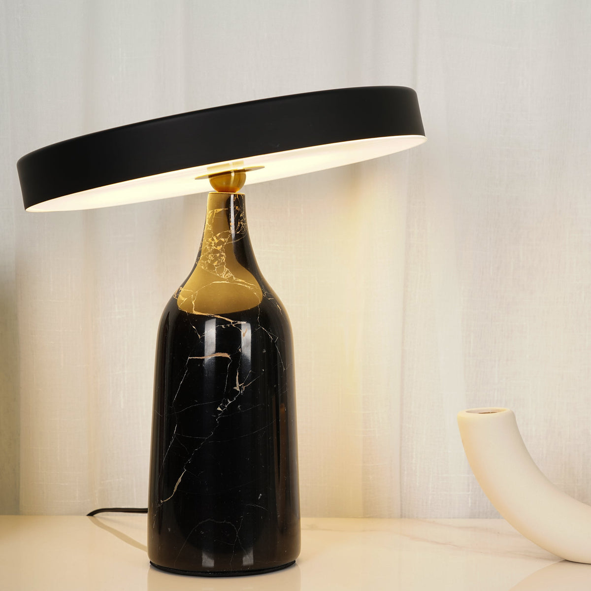 Black marble Clérachic table lamp lit warm in an ambient room setting, disc shade level beside a cream vase.