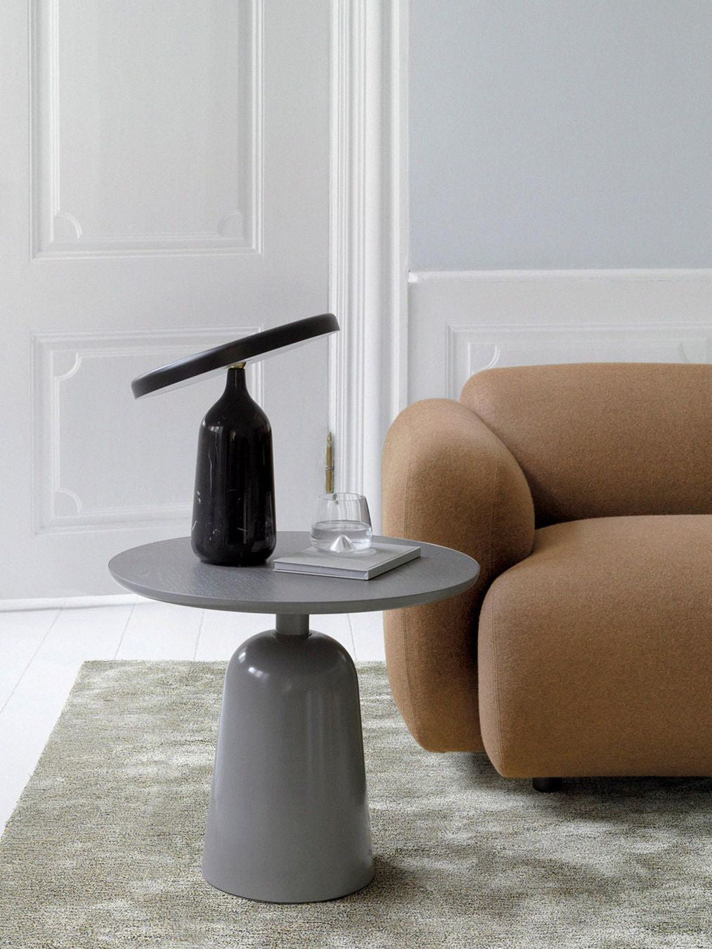 A round-top pedestal side table from the Clérachic collection, styled with a dark lamp-like vase beside a tan sofa.