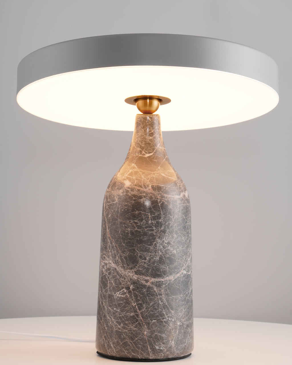 Grey marble Clérachic table lamp lit warm, photographed closer to show the glow beneath the disc shade.