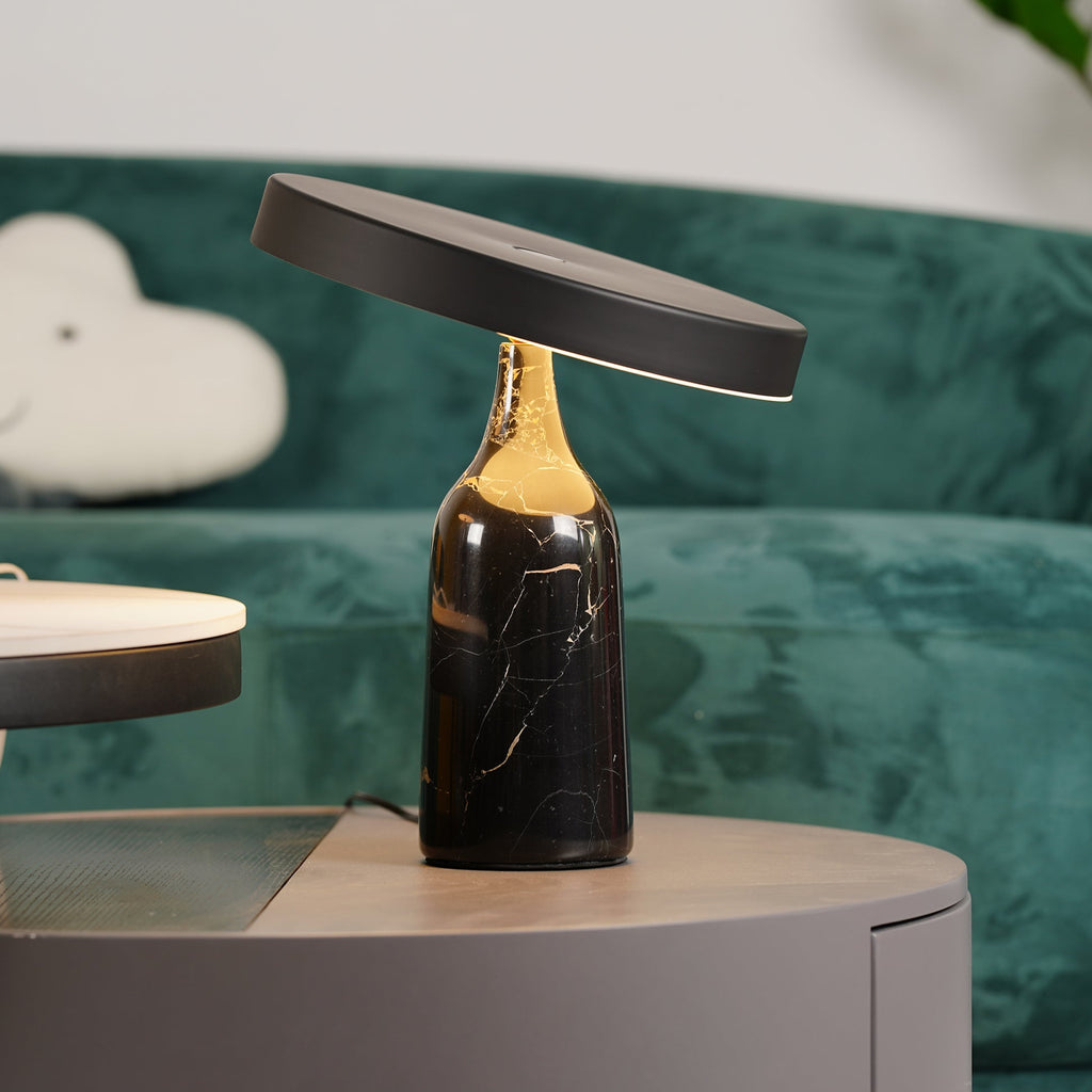 Close crop of the lit black marble Clérachic table lamp, disc shade tilted steeply on a dark green sofa.