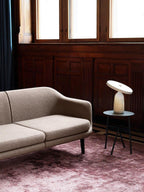 White marble Clérachic table lamp on a round metal side table, in a dark wood-panelled room with a beige sofa.