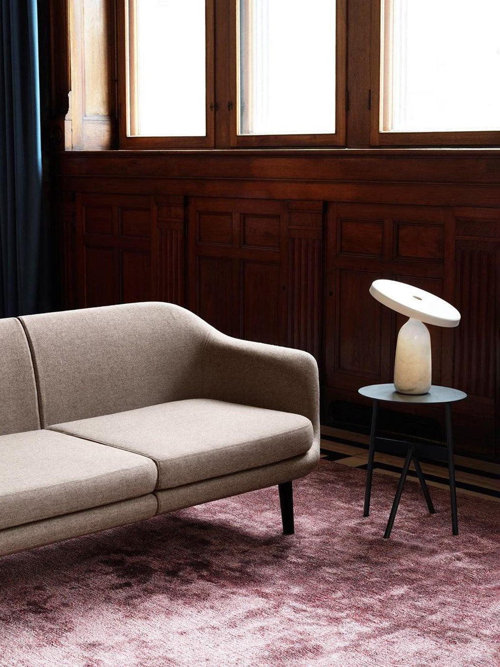 White marble Clérachic table lamp on a round metal side table, in a dark wood-panelled room with a beige sofa.