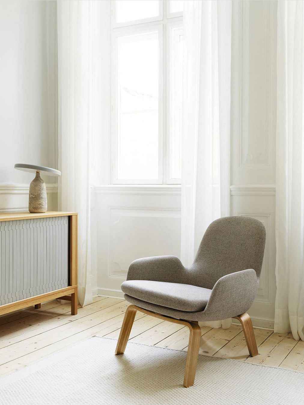 White marble Clérachic table lamp on a wooden console beside a radiator, grey armchair catching the daylight.