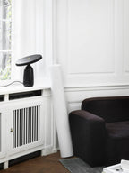 Black Clérachic table lamp on the windowsill, a wider view including the dark brown armchair and patterned rug.