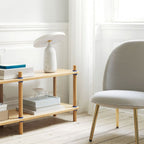 White marble Clérachic table lamp on the top tier of a wooden shelf stacked with books, beside a grey armchair.