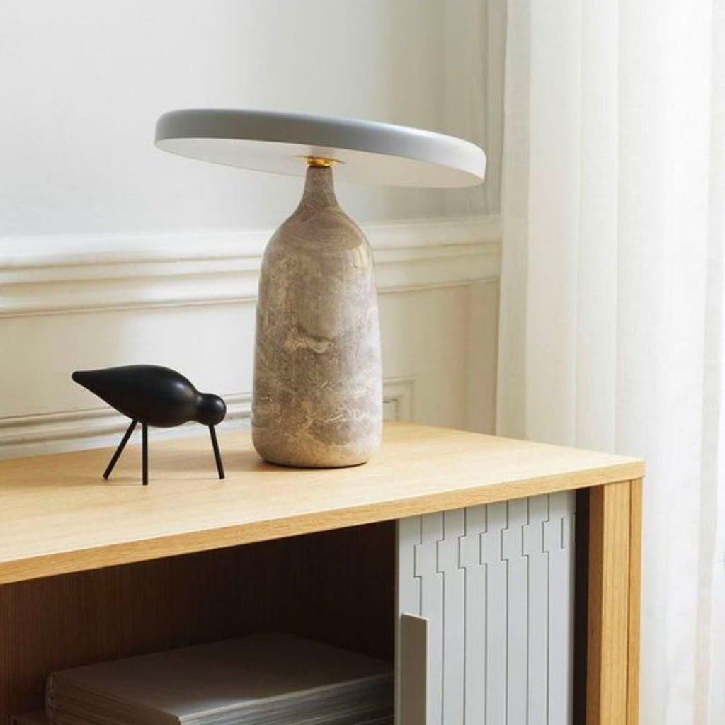 A pale marble Clérachic table lamp stands on a wooden shelf beside a small black bird figurine, near a window and radiator.
