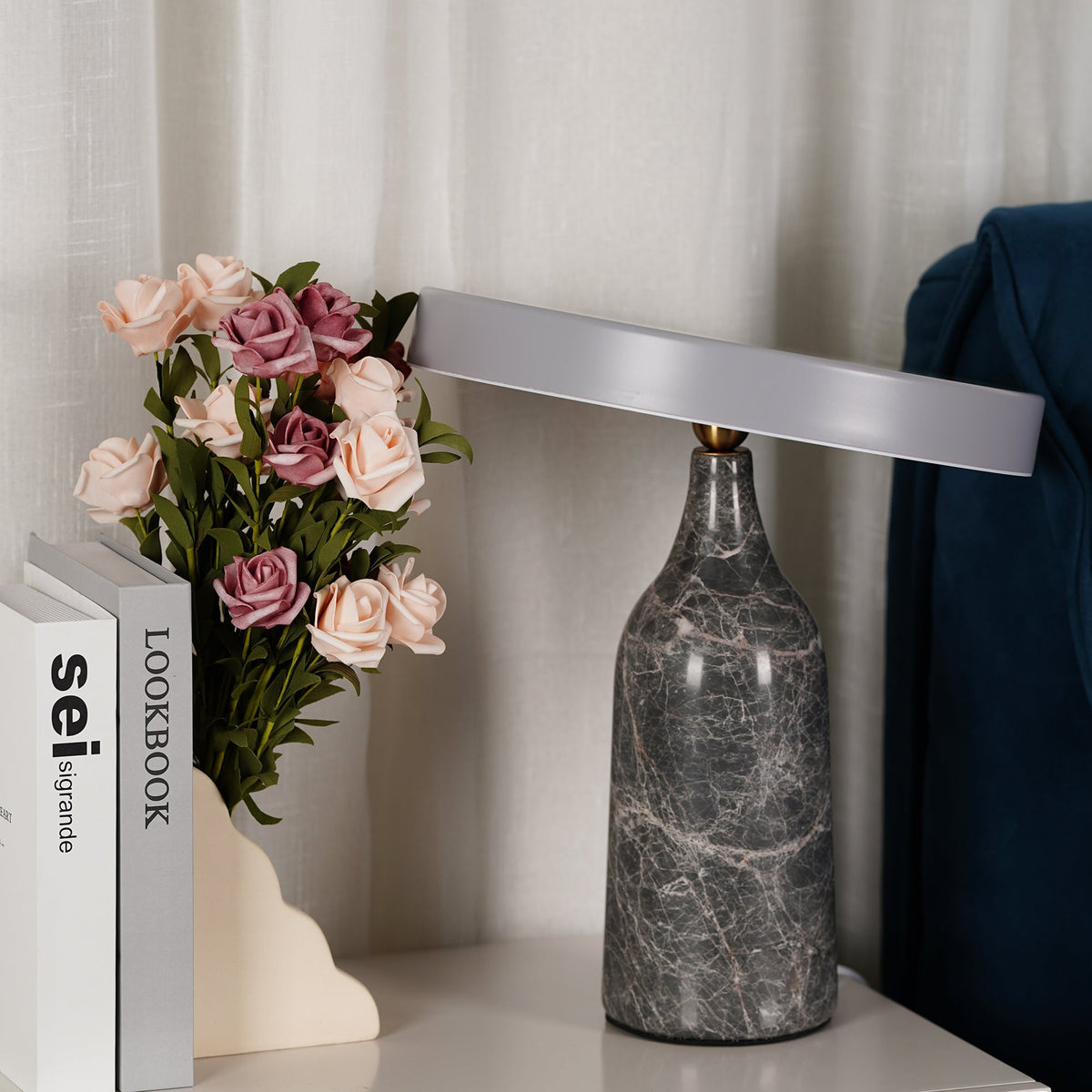 Grey marble Clérachic table lamp with its disc shade turned to a steep vertical angle, beside pink roses and books.
