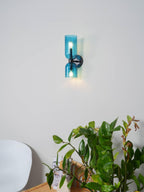 Beside a leafy green plant, this matt black Clérachic sconce shows pale blue glass shades on a white wall.
