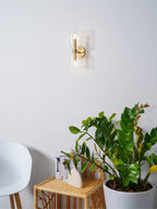 Potted plants sit beside this gold Clérachic wall light, its clear glass shades unlit on white.