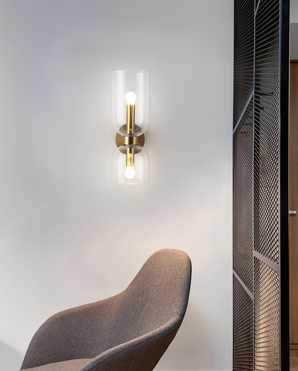 Beside a mesh room divider and a brown chair, this gold Clérachic wall light's clear shade glows.