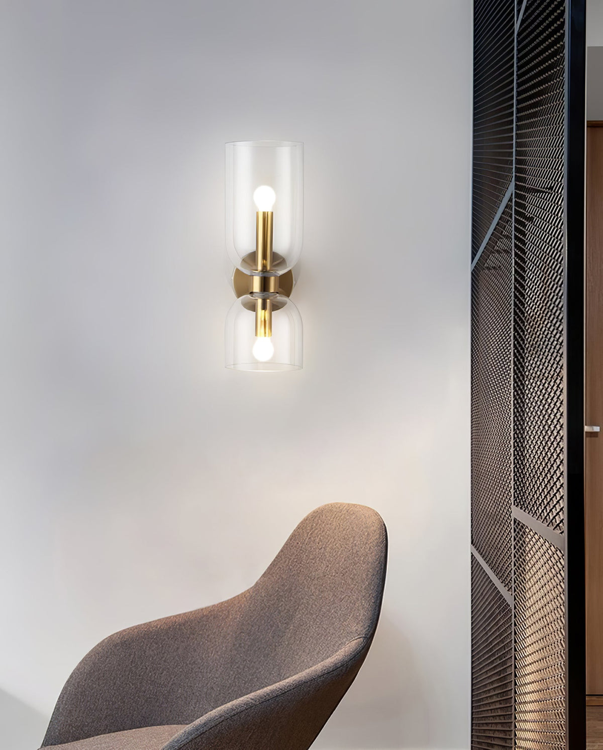 Beside a mesh room divider and a brown chair, this gold Clérachic wall light's clear shade glows.