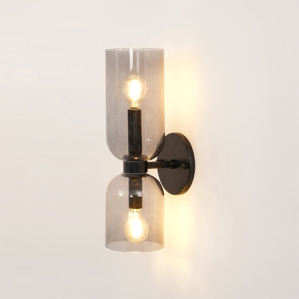 Clear glass cylinders expose the bulbs of this matt black Clérachic sconce, lit on a cream wall.