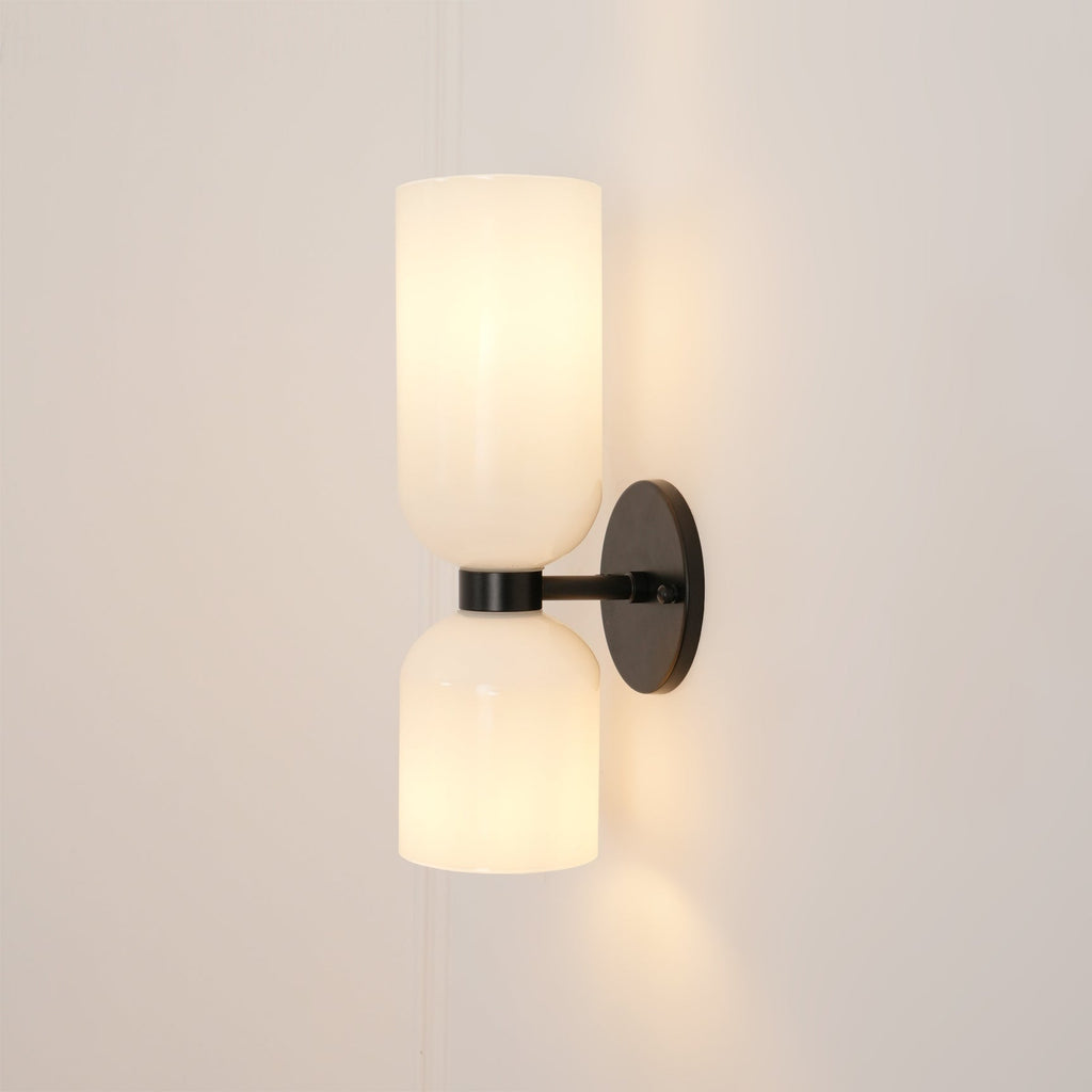 This Clérachic wall sconce in matt black holds two opal glass shades glowing softly against a warm taupe wall.