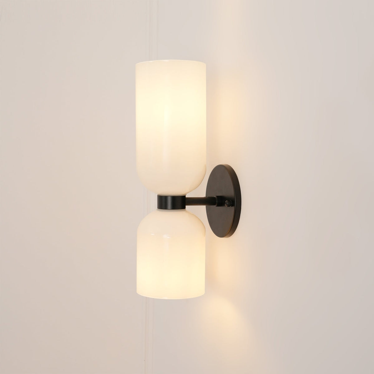This Clérachic wall sconce in matt black holds two opal glass shades glowing softly against a warm taupe wall.