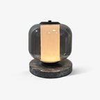 This Clérachic smoke-grey glass table lamp sits alone against a plain grey backdrop, its dark marble base and warm glow visible.