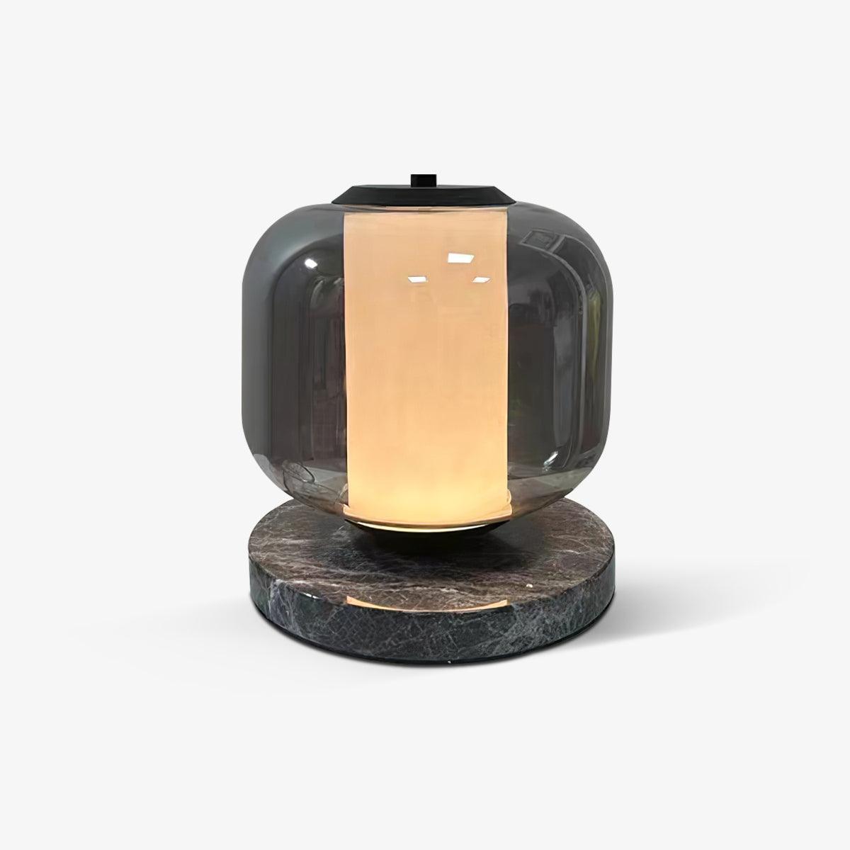 This Clérachic smoke-grey glass table lamp sits alone against a plain grey backdrop, its dark marble base and warm glow visible.