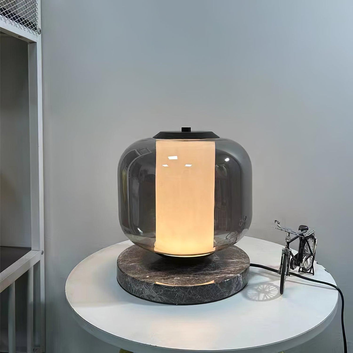 The Clérachic table lamp rests lit on a round bedside table, its cord trailing past a small toy bicycle behind it.