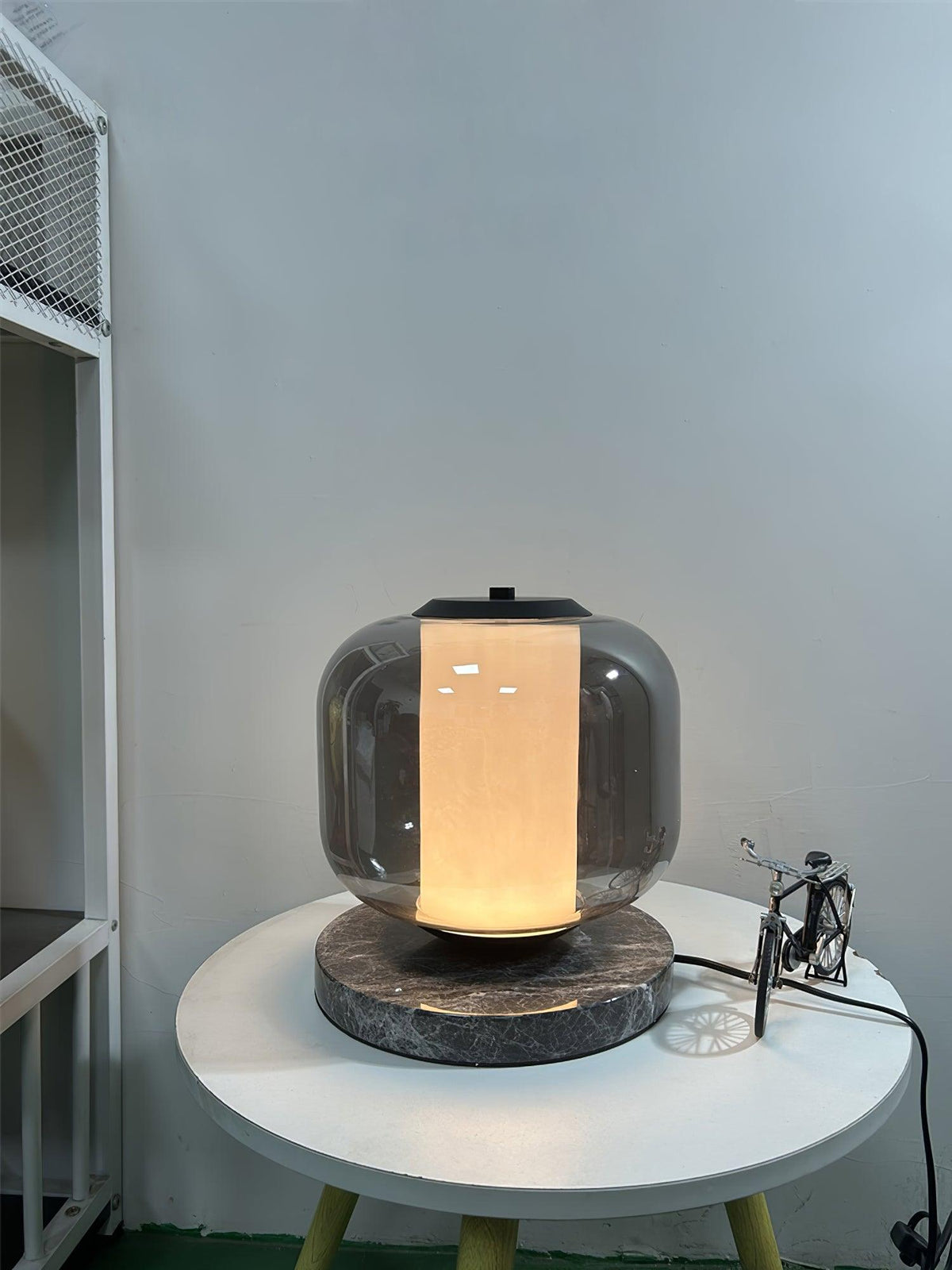 A tilted view of the Clérachic table lamp on its bedside table, with a metal shelving unit blurred in the background.