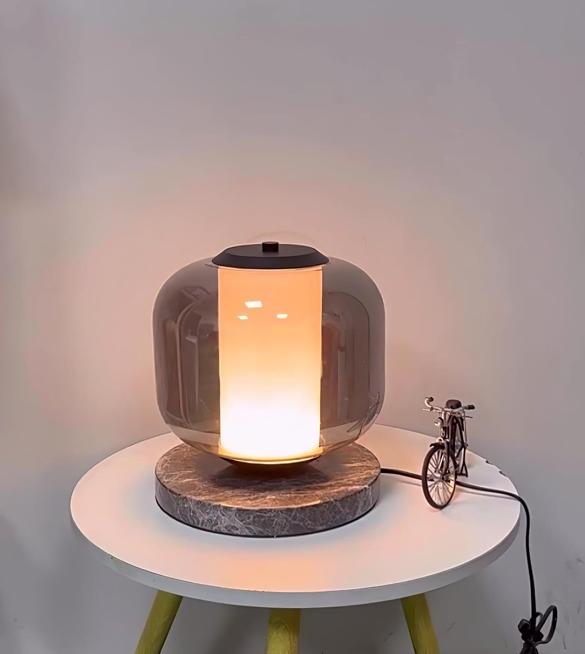 A closer angle on the lit Clérachic table lamp, its warm glass column glowing against a plain grey wall above the table.