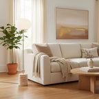 In a neutral modern living room, the Clérachic floor lamp sits near a fiddle-leaf fig plant beside a beige sofa.