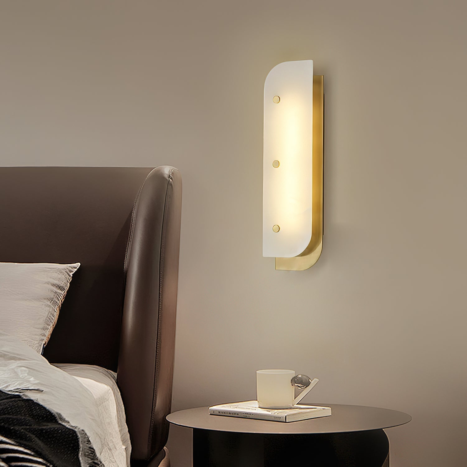Elaris Alabaster Wall Light in brass beside a leather headboard, lighting a bedside table with a cup and book.