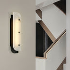 Tall Elaris Alabaster Wall Light in black on a stairwell wall, lit beside a brass-railed staircase.