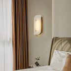 Short Elaris Alabaster Wall Light in brass by a curtained bedroom window, lit above a small nightstand.